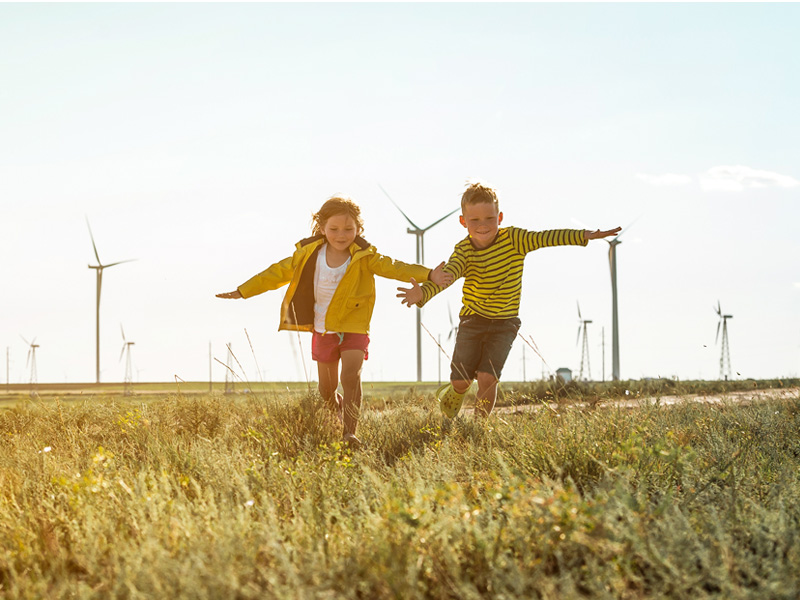 Zwei Kinder laufen über eine grüne Wiese, im Hintergrund stehen Windkraftanlagen